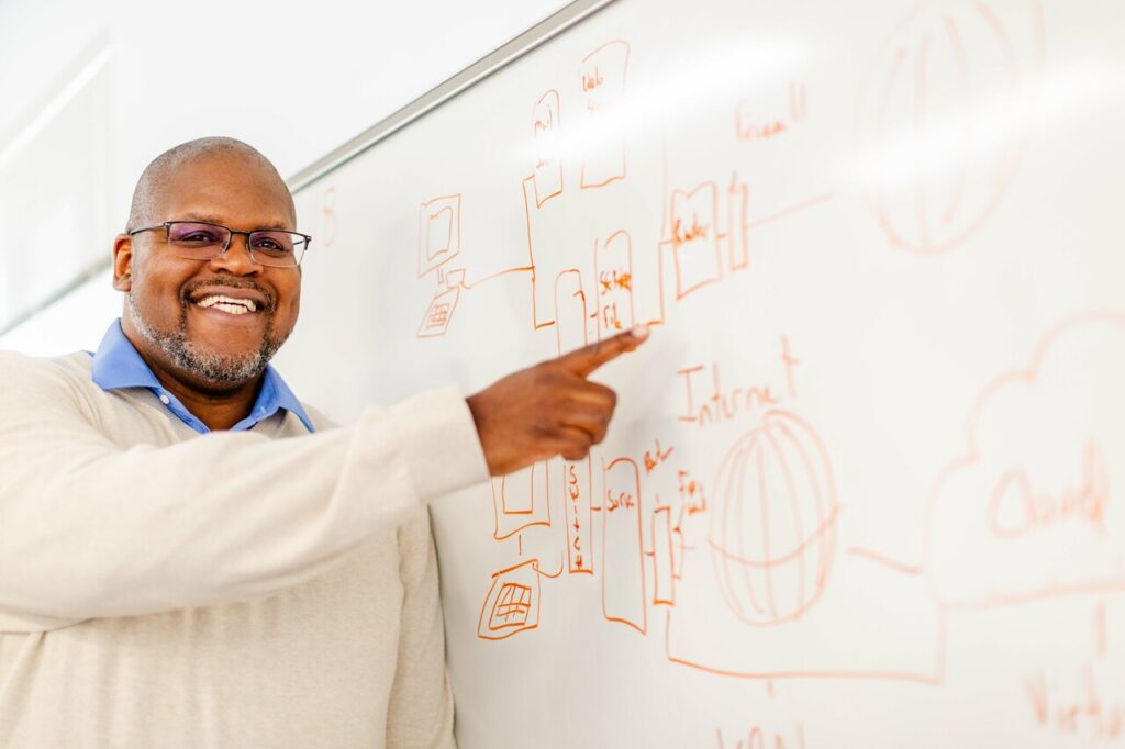 A smiling man with glasses points to a network architecture diagram drawn in orange marker on a whiteboard.