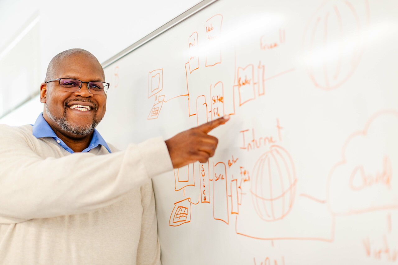 A smiling man with glasses points to a network architecture diagram drawn in orange marker on a whiteboard.